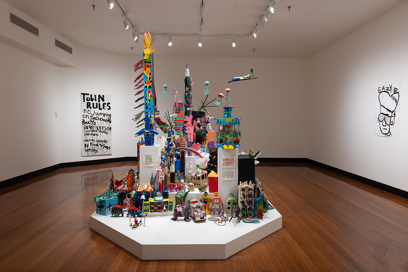 An image of a gallery space with a colourful sculpture of a city made from recycled materials. 
